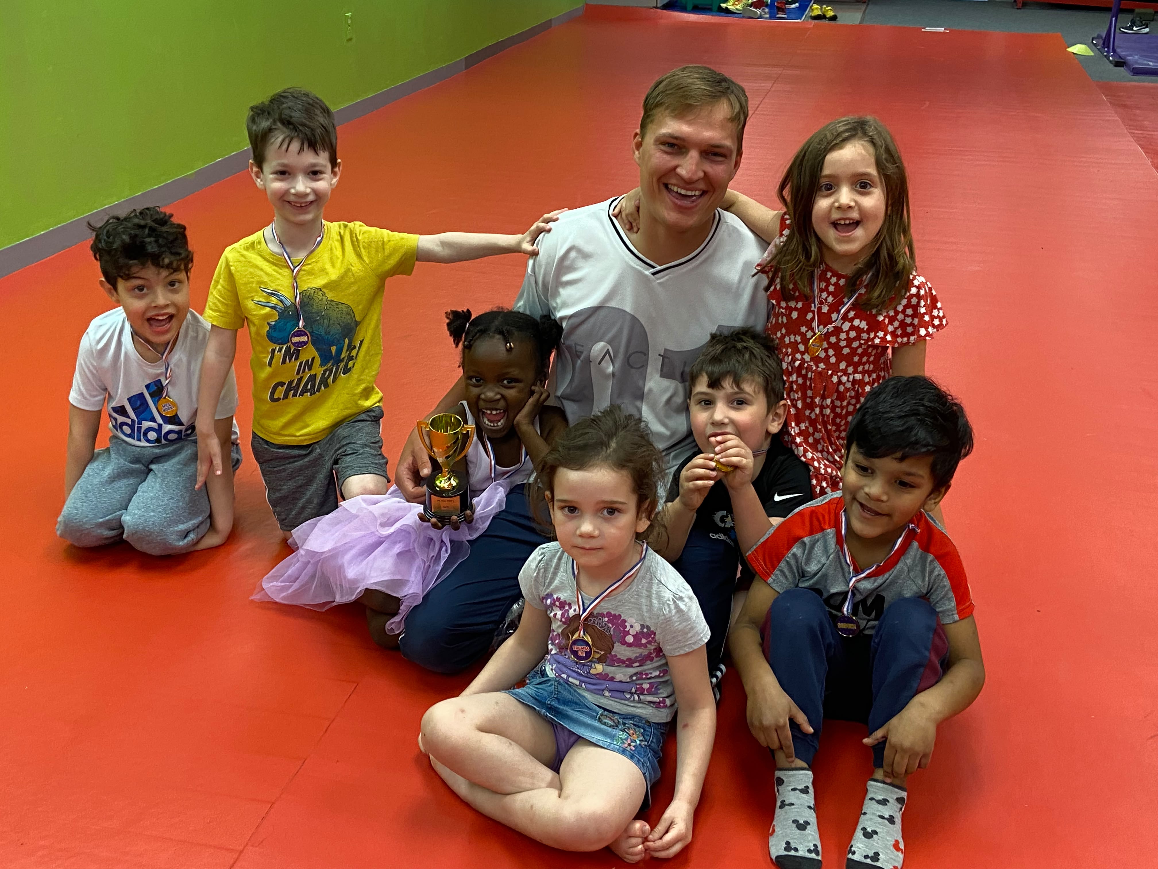 Children celebrating a gym achievement with their teacher, medals, and a trophy